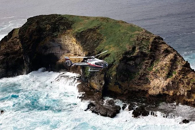 Ein Hubschrauber fliegt neben einem grünen Felsturm im Meer über weißer Brandung